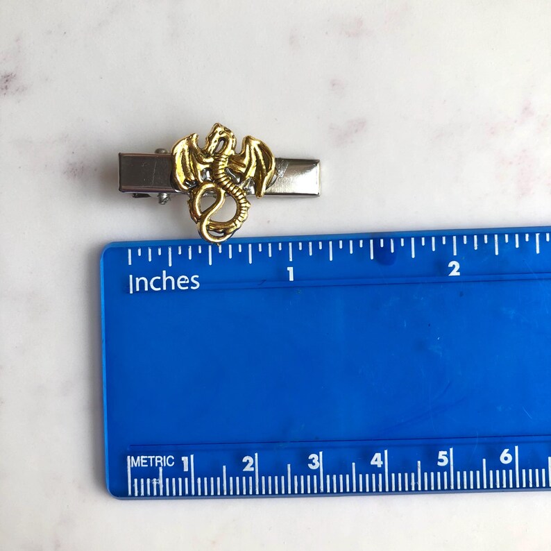 May include: A gold dragon tie clip with a silver clip. The dragon is approximately 2.5 cm long. The clip is on a white surface with a blue ruler that has both inches and metric measurements.