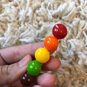 以下が含まれることがあります： 6つの丸いカラフルなビーズが付いた金色のヘアクリップ。ビーズは赤、オレンジ、黄色、緑、茶色、赤です。