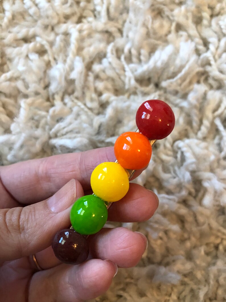 以下が含まれることがあります： 6つの丸いカラフルなビーズが付いた金色のヘアクリップ。ビーズは赤、オレンジ、黄色、緑、茶色、赤です。