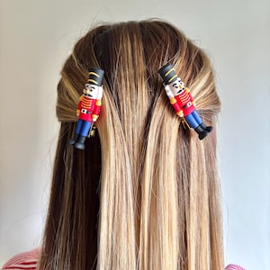 May include: Two toy soldier hair clips, each featuring a black hat, red jacket, blue trousers, and black boots. The clips are positioned in a person's hair, a blend of blonde and brown. The person is wearing a red and white striped top.