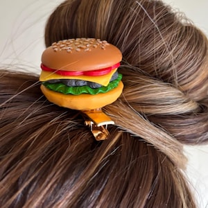 May include: A novelty hair clip shaped like a miniature hamburger. The clip features a sesame seed bun, a red tomato, yellow cheese, a black patty, and green lettuce. The clip is attached to a gold-colored metal clasp, designed to secure the clip to the hair.