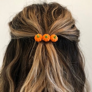 May include: Three small orange pumpkin hair clips with green stems. The clips are in a person's hair.