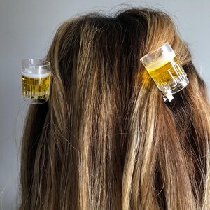 May include: Two clear plastic shot glass hair clips with a yellow liquid inside. The clips are in a person's hair.