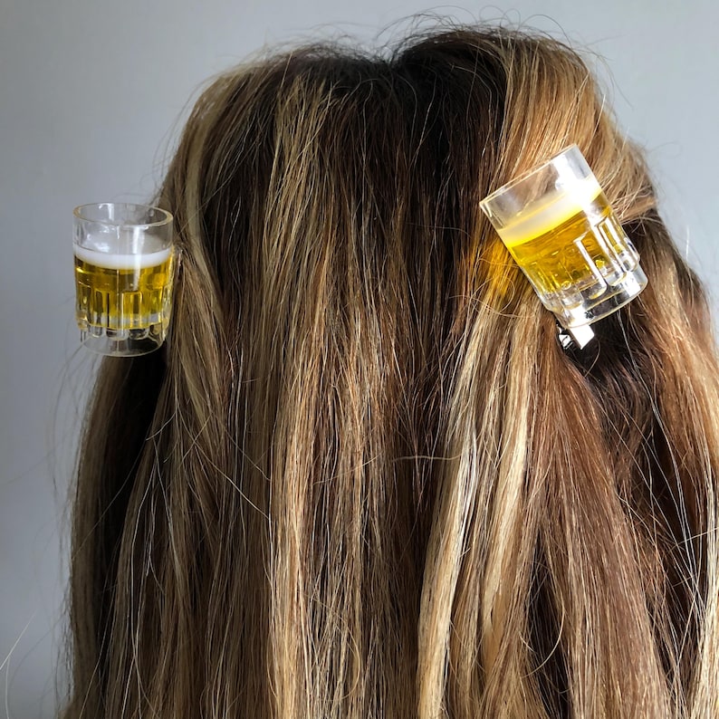 May include: Two clear plastic shot glass hair clips with a yellow liquid inside. The clips are in a person's hair.