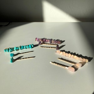 May include: Four gold hair clips with different coloured gemstone accents. The clips are arranged on a white surface. The gemstones are turquoise, amethyst, and rose quartz.