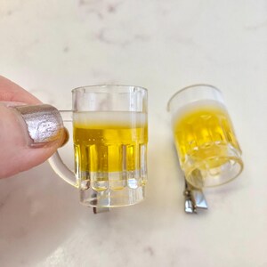 May include: Two miniature plastic beer mugs with a yellow liquid inside. The mugs are clear and have a frosted rim. The mugs are on a white surface.