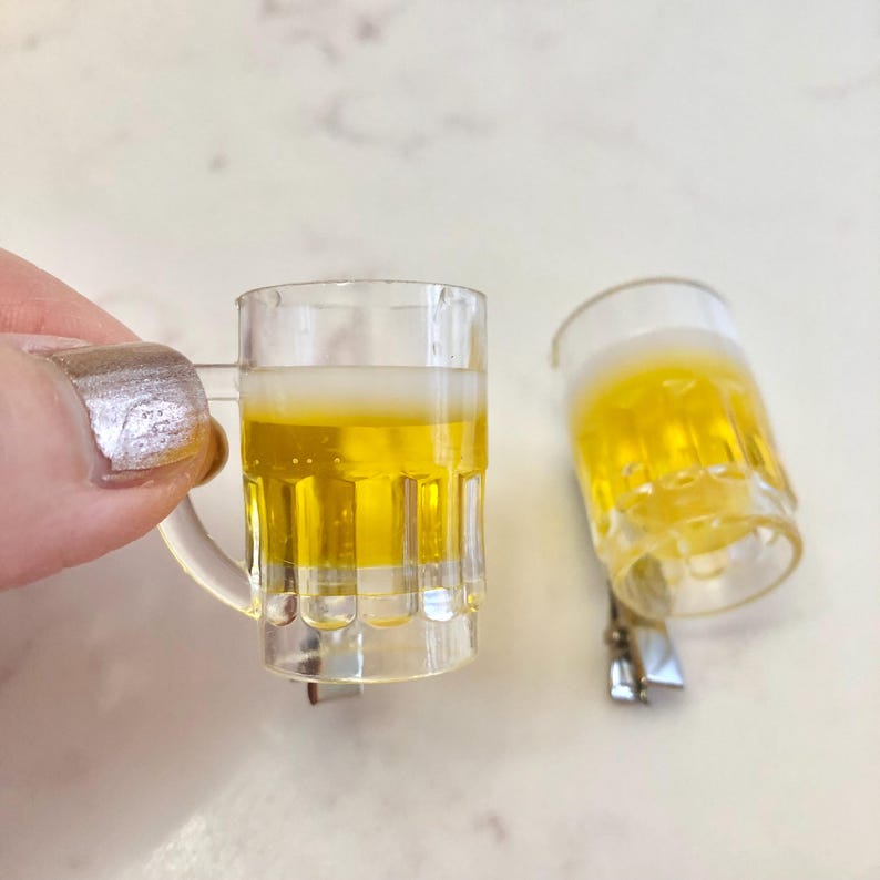 May include: Two miniature plastic beer mugs with a yellow liquid inside. The mugs are clear and have a frosted rim. The mugs are on a white surface.