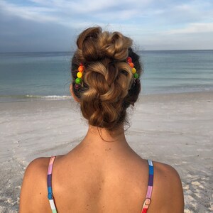 以下が含まれることがあります： カラフルなビーズのヘアクリップで編まれたお団子ヘアにした、長い茶色の髪の女性。彼女はレインボーストライプのビキニトップを着て、海を背景に砂浜に立っています。