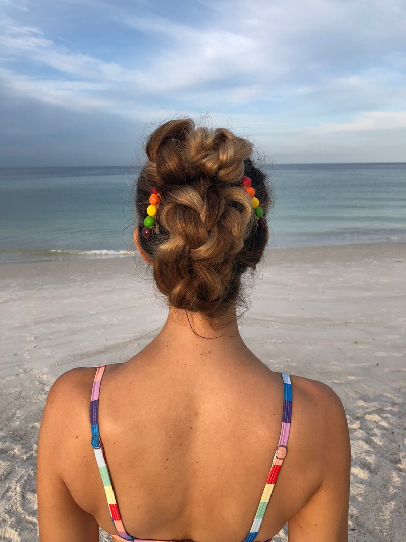 以下が含まれることがあります： カラフルなビーズのヘアクリップで編まれたお団子ヘアにした、長い茶色の髪の女性。彼女はレインボーストライプのビキニトップを着て、海を背景に砂浜に立っています。
