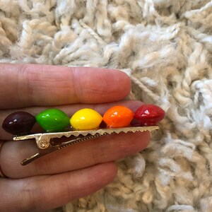 以下が含まれることがあります： 5つの丸いカラフルなビーズが付いた金色のヘアクリップ。ビーズは茶色、緑、黄色、オレンジ、赤です。