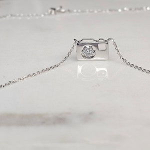 May include: A silver necklace featuring a miniature camera pendant. The camera pendant has a round, clear gemstone in the center, and is suspended from a delicate chain. The necklace is displayed on a white surface.