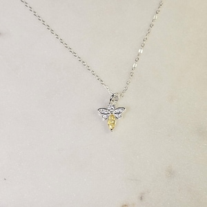 May include: A silver-toned necklace featuring a bee pendant. The bee pendant has a yellow, marquise-cut gemstone body, with white gemstones on the wings. The necklace chain is delicate and hangs on a white marble surface.