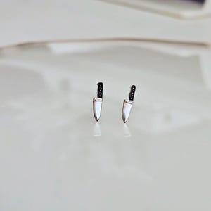 Puede incluir: Un par de pendientes plateados con forma de cuchillo y mangos negros. Los pendientes son pequeños y tienen un acabado pulido, que refleja la luz. Las hojas son lisas y los mangos tienen un aspecto texturizado.