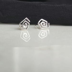 May include: A pair of silver stud earrings. Each earring has a geometric, spiral design within a pentagonal shape. The earrings are shown on a reflective surface with a dark fabric in the background.