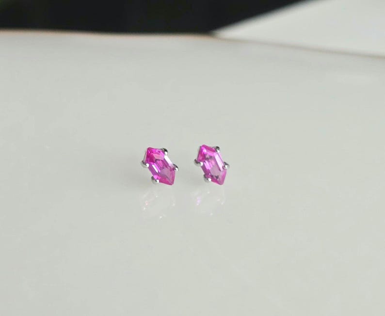 以下が含まれることがあります： 明るいピンク色のマーキス型宝石を銀色の金属にセットした、小さなスタッドピアス。白い表面に展示されており、背景はソフトフォーカスになっています。