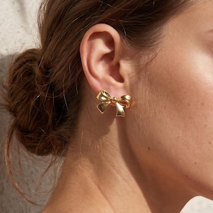 May include: A close-up of a person's ear with a gold bow-shaped earring. The earring is a solid, shiny gold color and has a detailed bow design. The person's skin is fair, and their hair is pulled back.