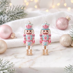 May include: A pair of stud earrings shaped like nutcracker soldiers. Each earring features a white, pink, and light blue enamel design with gold-tone accents, a small white pearl-like bead at the top, and a post-back closure mechanism.