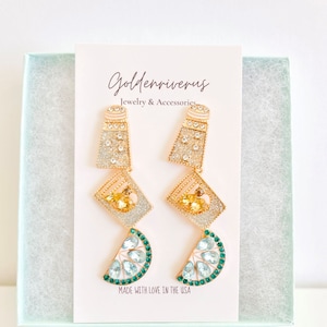 May include: A pair of gold-toned earrings with a unique design. The earrings feature a salt shaker shape, diamond-shaped center, and a lime slice at the bottom, all adorned with sparkling crystals. The earrings are displayed on a card that reads "Goldenriverus Jewelry & Accessories."