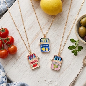 May include: Three gold-tone chain necklaces featuring rectangular sardine tin pendants. One pendant is blue with lemon graphics, one is cream with tomato graphics, and one is pink with olive graphics. Each pendant includes a small metal key detail.