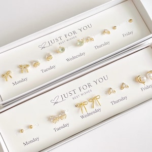 May include: Two white rectangular gift boxes containing gold-toned stud earrings arranged by day of the week from Monday to Friday. The boxes feature the text JUST FOR YOU BEST WISHES. The earrings include designs like bows, pearls, and crystals.