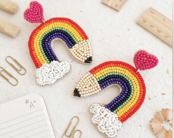 May include: A pair of beaded rainbow earrings with a pencil design. The earrings feature a red heart at the top, a rainbow arch, a white cloud, and a pencil tip. The earrings are colorful and whimsical.