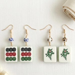May include: Four pairs of gold-toned earrings. Each earring features a white rectangular tile with a design, connected to a decorative bead and a gold-colored hook. Two pairs have circle designs, and two pairs have a green dragon design.