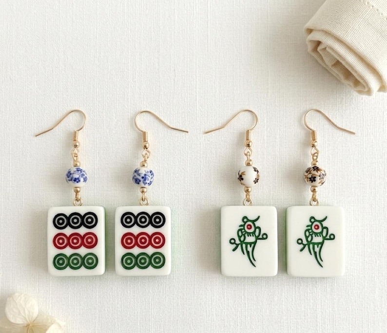 May include: Four pairs of gold-toned earrings. Each earring features a white rectangular tile with a design, connected to a decorative bead and a gold-colored hook. Two pairs have circle designs, and two pairs have a green dragon design.