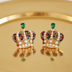 May include: A pair of gold-toned crown earrings with colorful gemstones and faux pearls. Each earring features a teardrop-shaped green gem at the top. The earrings are displayed on a gold-colored surface.