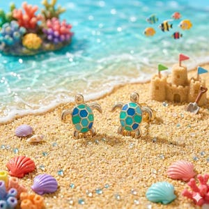 May include: Two gold-toned turtle rings with blue and green enamel shells rest on a sandy beach. The scene includes colorful seashells, coral, and a miniature sandcastle with flags, evoking a coastal theme.