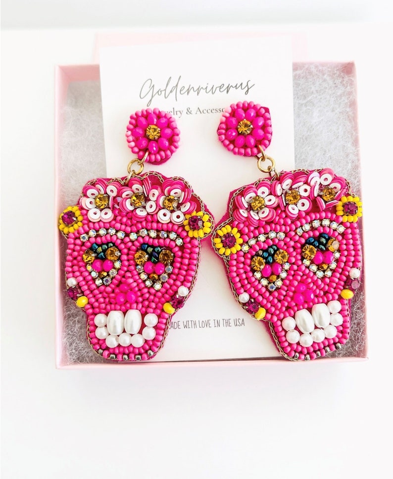 May include: A pair of hot pink beaded skull earrings with floral accents in a pink gift box. The earrings feature white pearl teeth and are embellished with yellow, gold, and purple beads. The card in the box reads "Goldenriverus Jewelry & Accessories."