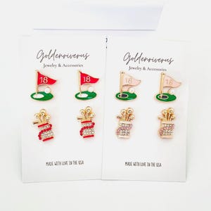 May include: Two sets of golf-themed earrings on white cards. Each set includes a golf flag with the number 18, a golf ball, and a golf bag with golf clubs. The earrings are gold-toned with red, green, and pink accents. The text "MADE WITH LOVE IN THE USA" is printed on the cards.