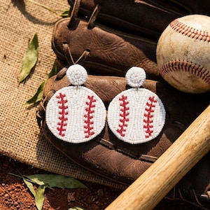 May include: A pair of round, white beaded earrings featuring a red stitched baseball pattern. Each earring consists of a small circular stud attached to a larger circular drop, mimicking the appearance of a classic baseball.