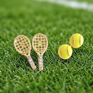 May include: Gold-coloured tennis racket earrings with clear crystal accents and two yellow tennis ball stud earrings. The earrings are set against a backdrop of green grass, creating a sporty theme.