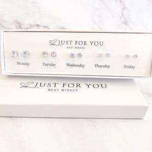 May include: A white box set of earrings with the text "JUST FOR YOU BEST WISHES". The set includes five pairs of earrings labeled Monday through Friday. Each pair features a different design, with clear, sparkling stones.