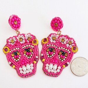 May include: Pair of hot pink beaded skull earrings. Each earring features a skull design with white, yellow, and gold accents. The earrings have a post back and are designed for pierced ears.