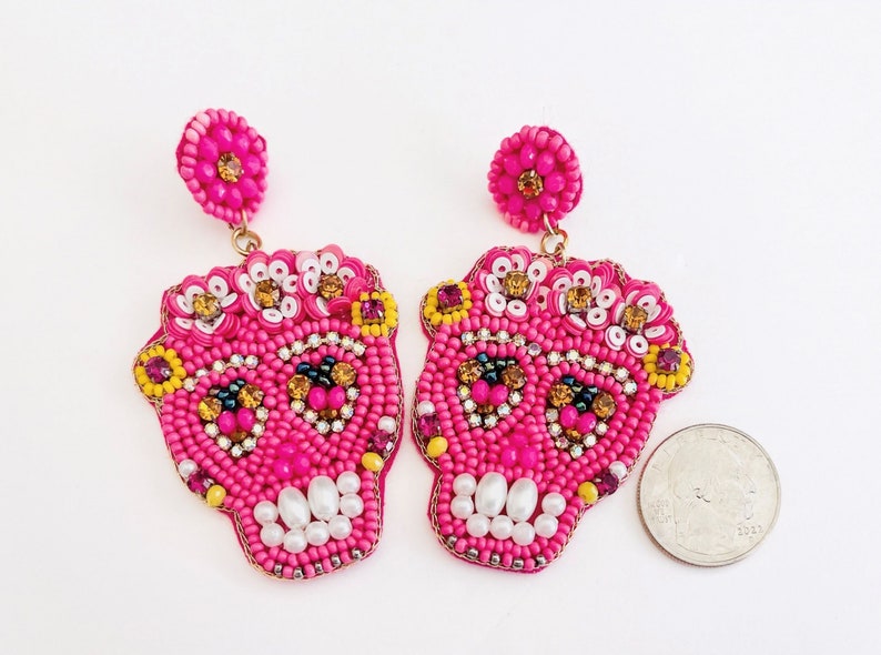 May include: Pair of hot pink beaded skull earrings. Each earring features a skull design with white, yellow, and gold accents. The earrings have a post back and are designed for pierced ears.