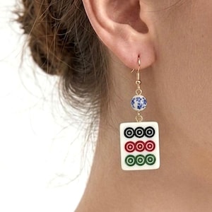 May include: Gold-tone dangle earrings featuring a white tile with three rows of three circles in black, red, and green. Each earring has a blue and white floral bead and a gold-tone hook.