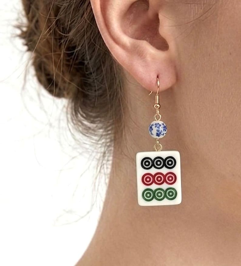 May include: Gold-tone dangle earrings featuring a white tile with three rows of three circles in black, red, and green. Each earring has a blue and white floral bead and a gold-tone hook.