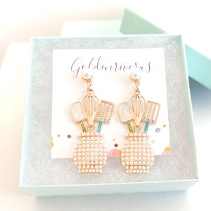 May include: Goldtone earrings with a pearl at the top and a design of a vase with kitchen utensils inside. The vase is made of white pearls and the utensils are goldtone with colorful rhinestones. The earrings are in a white box with the text "Goldenrivers" on a card.