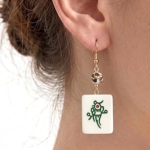 May include: Gold-tone dangle earring featuring a white rectangular tile with a green and red design, suspended from a floral bead and a fish hook. The earring is worn on the ear.