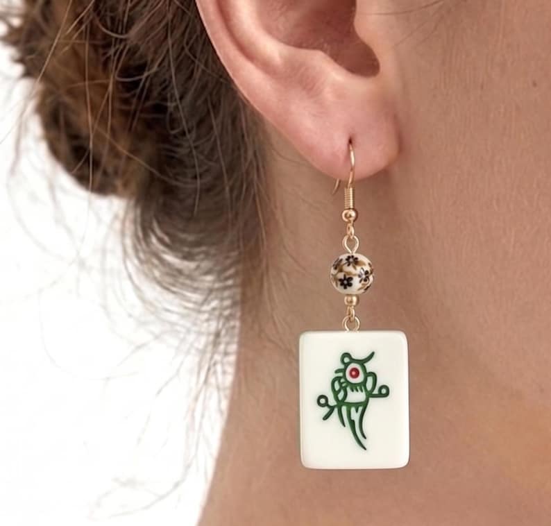 May include: Gold-tone dangle earring featuring a white rectangular tile with a green and red design, suspended from a floral bead and a fish hook. The earring is worn on the ear.