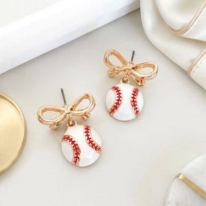 May include: A pair of gold-colored bow-shaped stud earrings. Each earring has a white baseball charm with red stitching. The earrings are displayed on a white surface with a gold-colored tray and white fabric in the background.