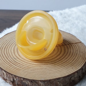 May include: A yellow silicone baby bottle nipple with a wide base and a small hole in the center. The nipple is sitting on a wooden surface.