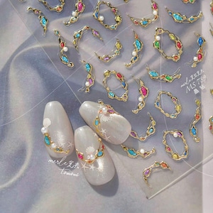 May include: A sheet of nail art stickers featuring gold-colored, jewel-toned embellishments. The designs include curved shapes, circles, and pearl accents. Three manicured nails are decorated with similar designs, including seashells and star details.