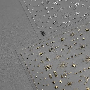 Peut inclure: Deux feuilles d'autocollants pour nail art avec des motifs célestes. Les autocollants comprennent des étoiles, des lunes et d'autres formes en argent et en or. Le texte "to 2020" est visible sur une feuille. Les autocollants sont sur un support transparent.