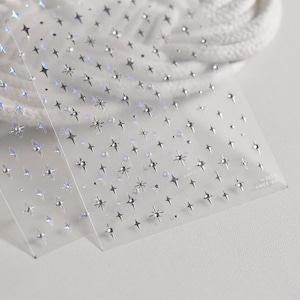 May include: Clear nail art stickers with various silver star designs. The stickers feature different star shapes and sizes, some with a gem-like centre. The stickers are on a clear backing.