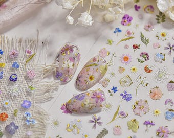 May include: Close-up of nail art featuring a variety of small, colorful flowers and gold foil accents. Three nails are decorated with floral designs. The image also shows a sheet of floral nail art stickers and dried flowers.