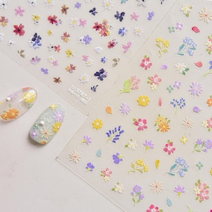 Könnte beinhalten: Blätter mit Nagelkunst-Aufklebern mit einer Vielzahl farbenfroher Blumendesigns. Die Aufkleber umfassen Gänseblümchen, Schmetterlinge und andere Blumenformen in Rosa, Gelb, Lila und Weiß. Zwei manikürte Nägel sind ebenfalls sichtbar.