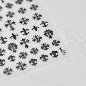 Könnte beinhalten: Bogen mit Nagelkunst-Aufklebern mit verschiedenen schwarzen dekorativen Designs, darunter Kreuze, Fleur-de-Lis und Blumenmuster. Die Aufkleber befinden sich auf einer transparenten Unterlage. In der Ecke des Bogens steht der Text "10 2104".