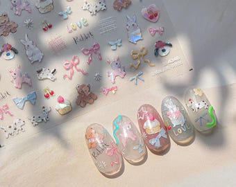 May include: A sheet of nail art stickers featuring various designs, including teddy bears, bunnies, bows, and cupcakes. The stickers are in pastel colors with glitter accents. Several manicured nails are also shown, each with a different design, including the word "Love".
